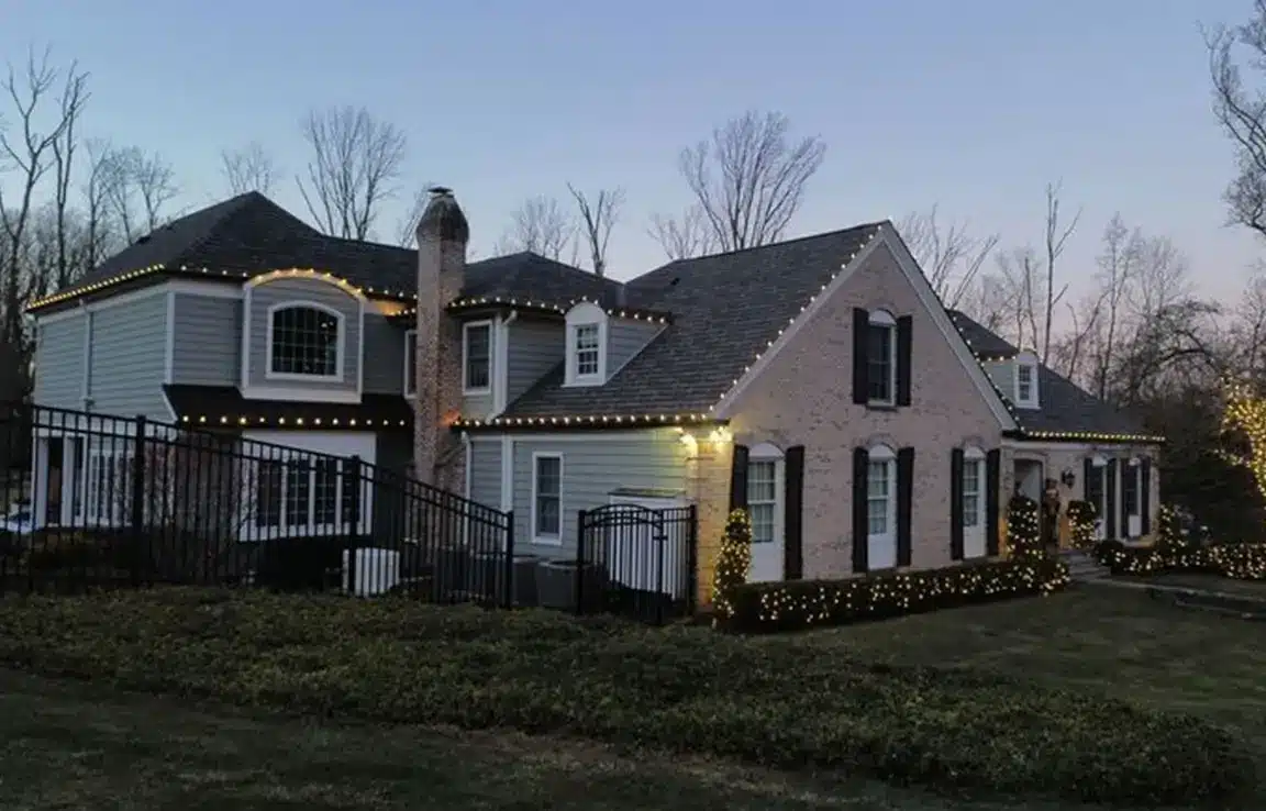 Large two-story suburban house at dusk with white string lights outlining the roof and bushes, brick and siding facade, chimney and leafless trees in background.
